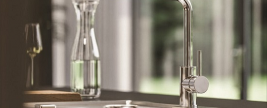 Elegante grifería de cocina cromada Alveus con líneas redondeadas, instalada en un fregadero en una cocina moderna con una vista acristalada de la vegetación.