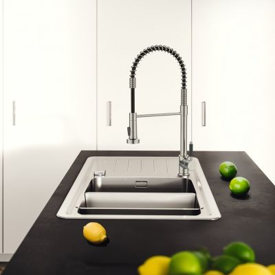 Grifo de cocina profesional con caño extraíble flexible de acero inoxidable, instalado junto a un fregadero negro en una cocina luminosa con elementos de madera y una ventana panorámica
