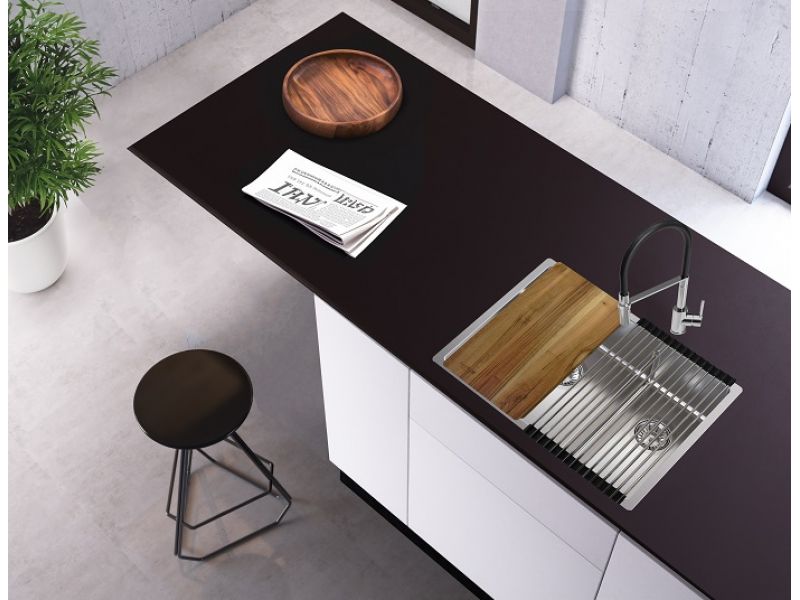 Vista desde arriba de una isla de cocina con armarios blancos y una encimera oscura en la que está integrado un fregadero de acero inoxidable con escurridor y tabla de cortar de madera; sobre la encimera hay una bandeja de madera y un periódico, y junto a la isla, un taburete alto y una planta en maceta.