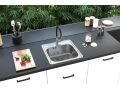 Cocina moderna con encimera negra mate y armarios blancos, en la que está integrado un fregadero cuadrado de acero inoxidable con un grifo alto negro; junto a él hay una tabla con verduras, utensilios de cocina y cuencos, y al fondo una pared de cristal con vistas a la vegetación.
