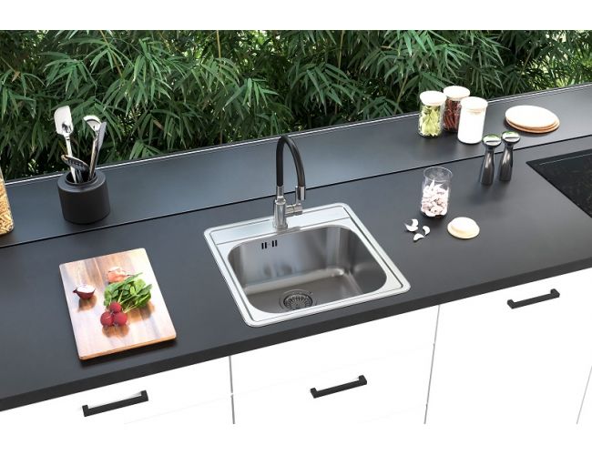 Cocina moderna con encimera negra mate y armarios blancos, en la que está integrado un fregadero cuadrado de acero inoxidable con un grifo alto negro; junto a él hay una tabla con verduras, utensilios de cocina y cuencos, y al fondo una pared de cristal con vistas a la vegetación.