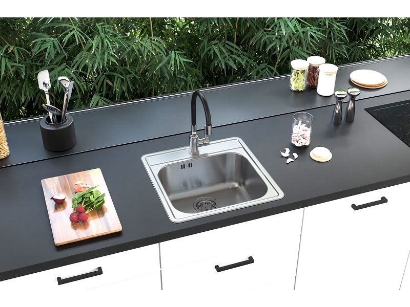Cocina moderna con encimera negra mate y armarios blancos, en la que está integrado un fregadero cuadrado de acero inoxidable con un grifo alto negro; junto a él hay una tabla con verduras, utensilios de cocina y cuencos, y al fondo una pared de cristal con vistas a la vegetación.