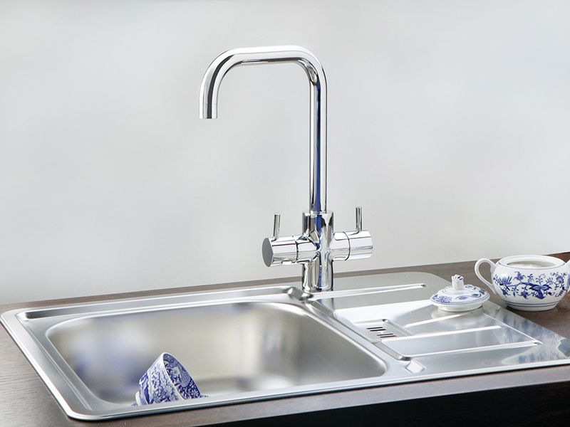 Inox kuhinjsko pomivalno korito z odcejalno površino in visoko kromirano armaturo; na odcejalni površini je skodelica s krožničkom in čajnikom z modrim vzorcem.